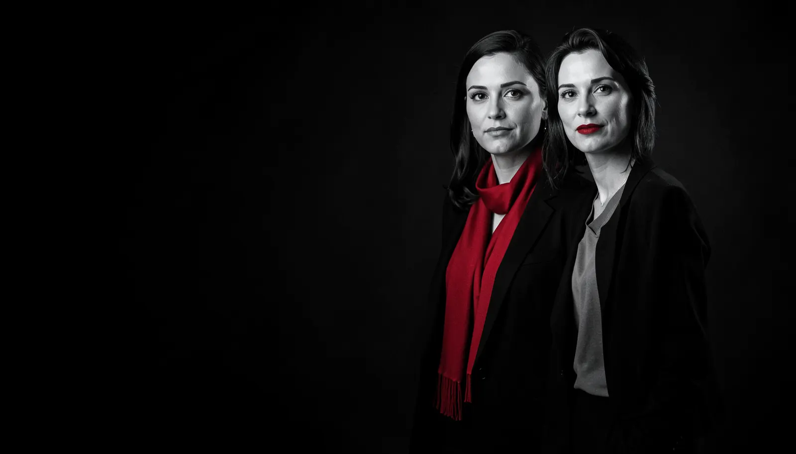Zwei selbstbewusste Frauen in schwarzen Blazern vor dunklem Hintergrund, das Bild ist schwarz-weiß, nur Schal und Lippenstift leuchten rot